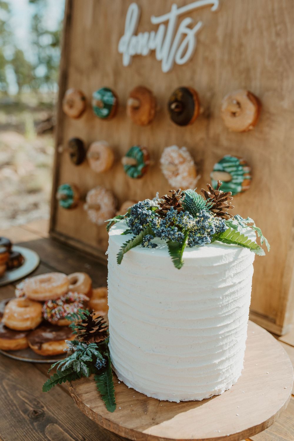 White wedding cake decorated with fresh greenery next to wooden donut wall display.