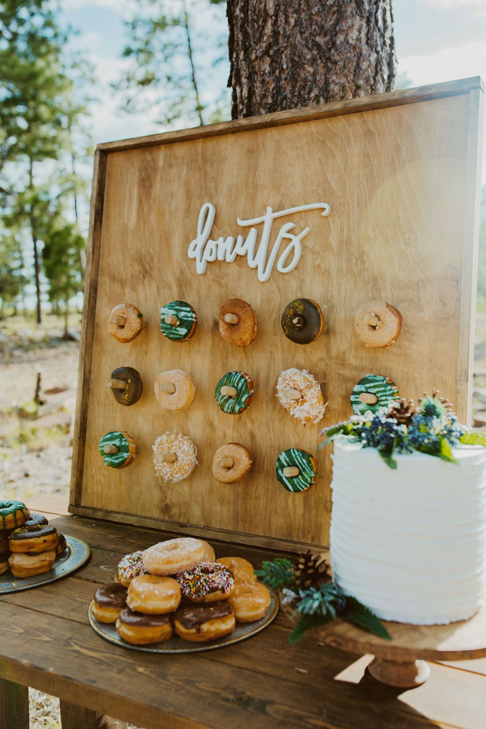 Wooden donut wall with turquoise and glazed donuts displayed next to white cake at outdoor event.