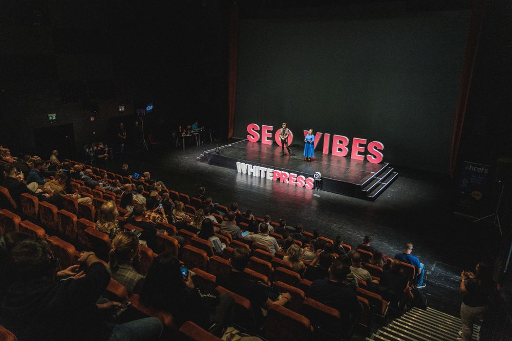 Audience in dark theater faces illuminated SEO VIBES sign on stage during conference presentation.