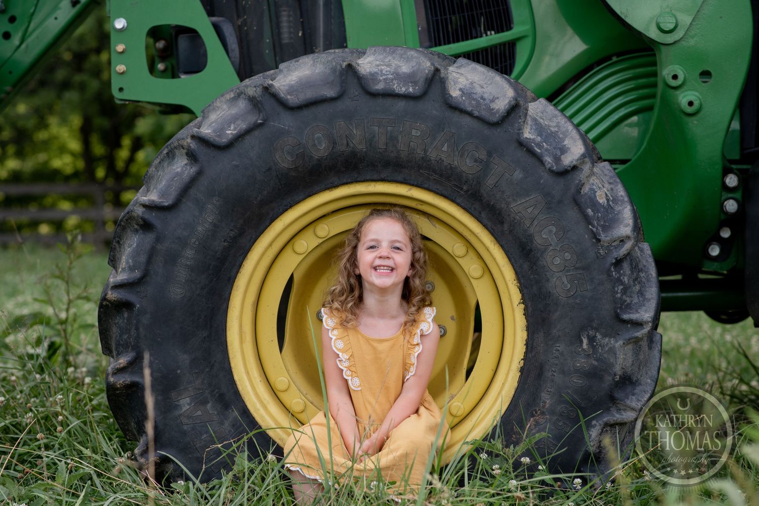 Young girl in yellow dress poses inside large tractor wheel on farm.