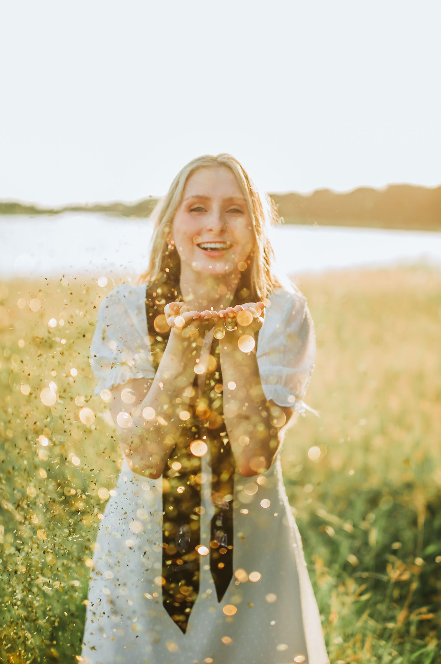 Person in white dress blowing glitter in golden sunlight at sunset.
