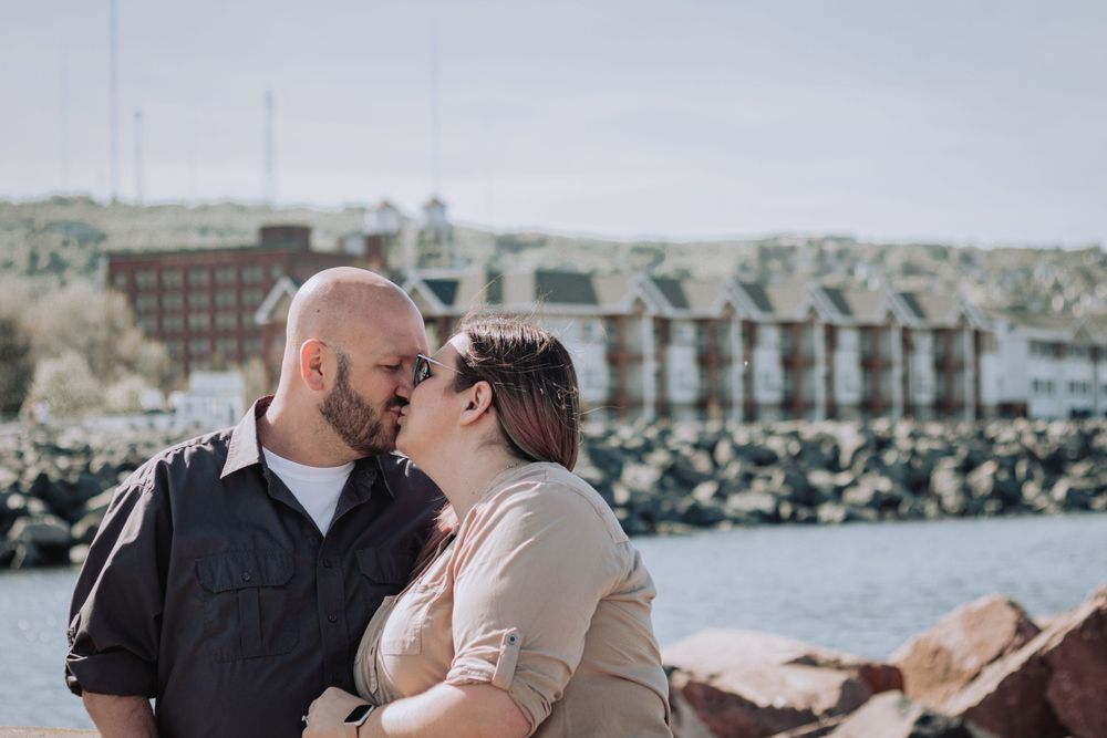 A couple shares a tender kiss against a waterfront backdrop with rocks and buildings in the distance.