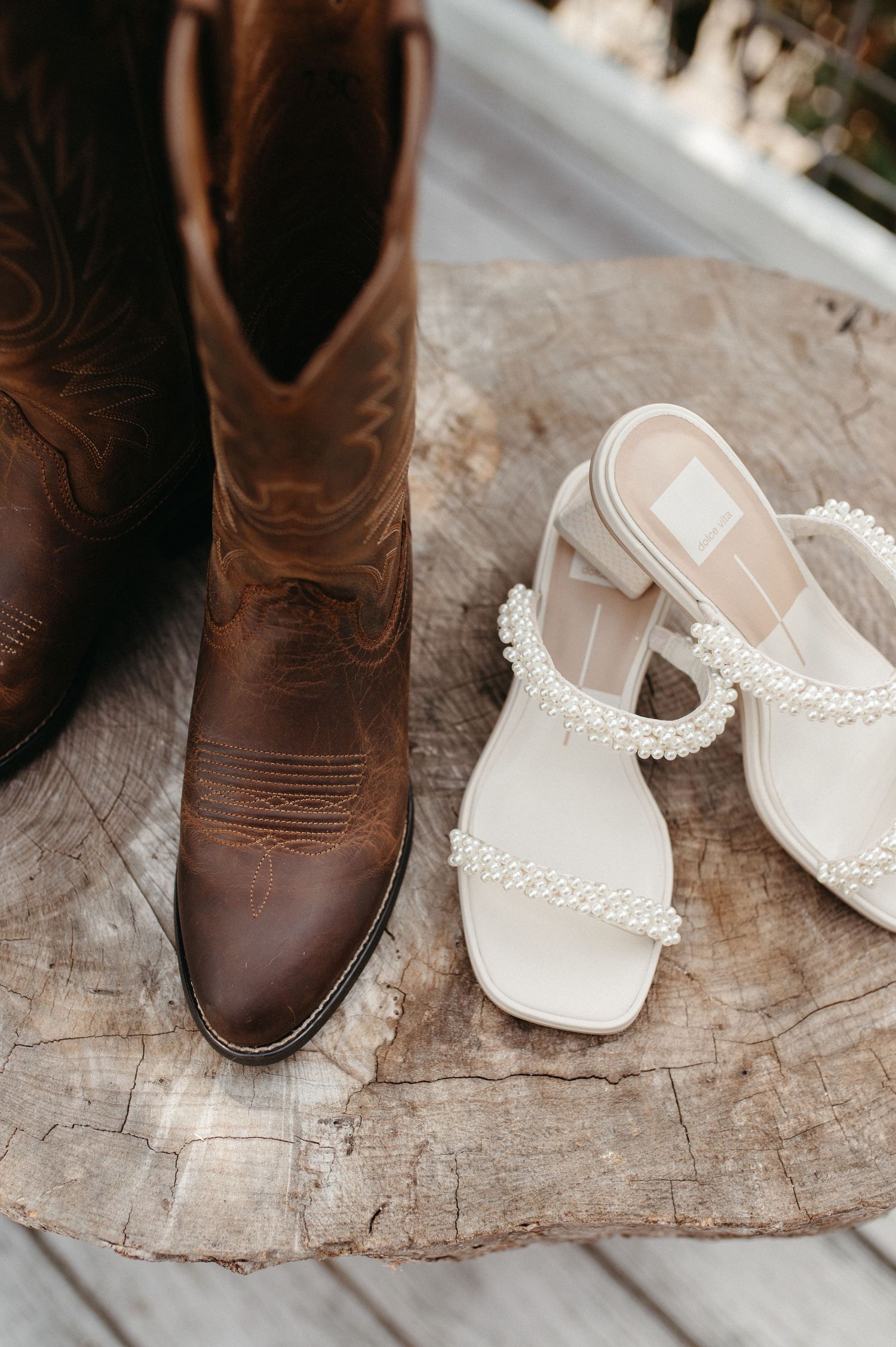 Brown leather cowboy boots and white pearl-embellished sandals arranged on a rustic wooden surface.