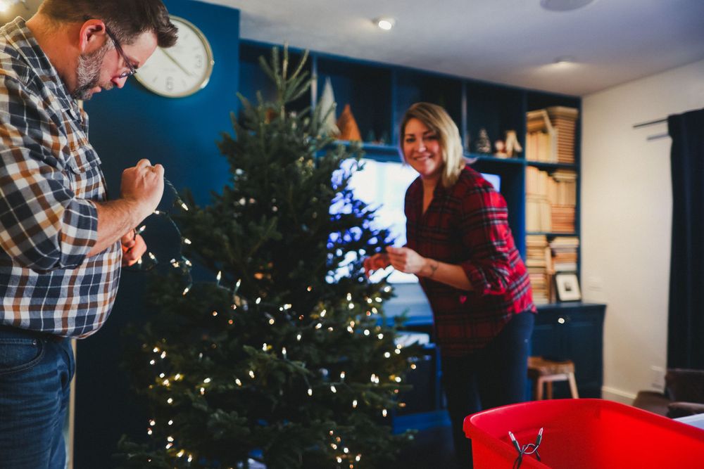 People decorating a tall Christmas tree in a living room with framed artwork.