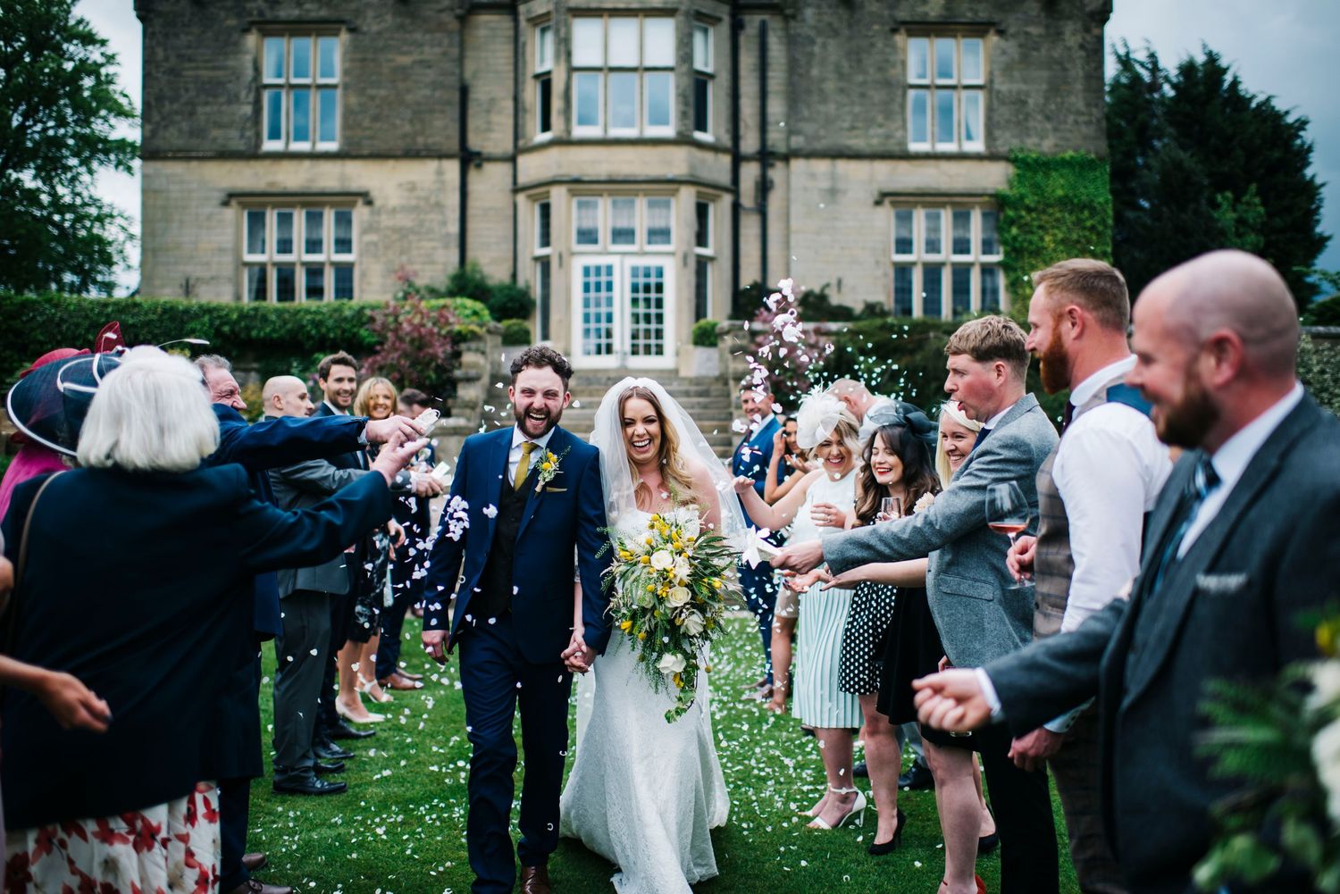 Wedding guests shower newlyweds with confetti outside a grand stone mansion during an outdoor celebration.