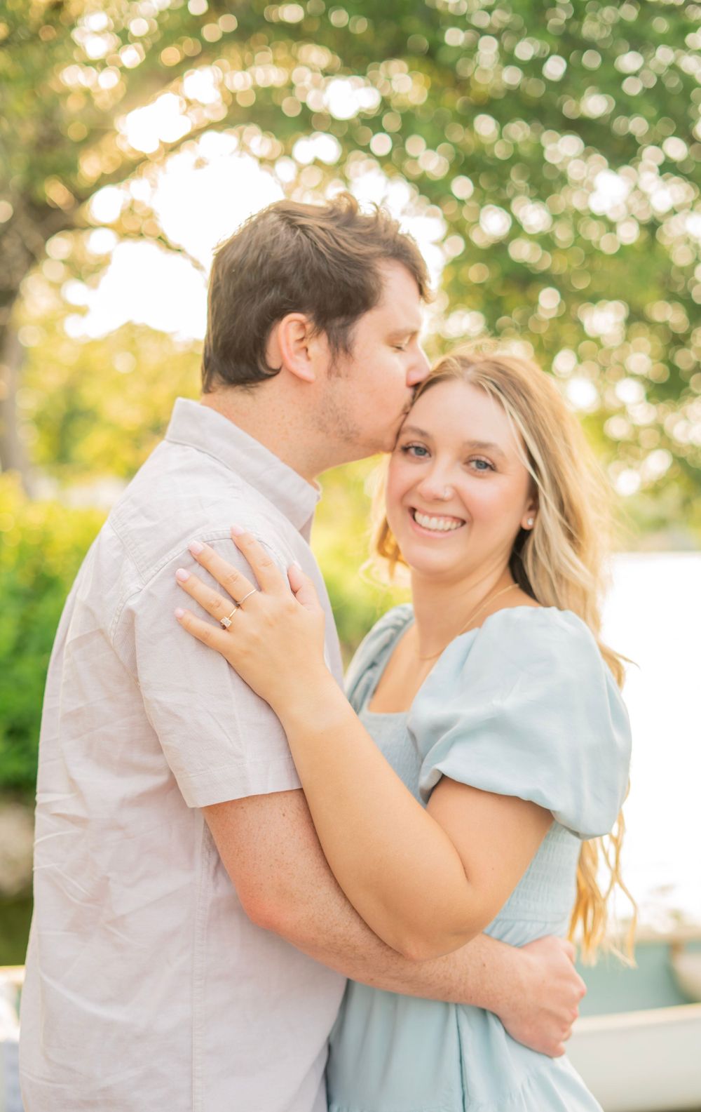 Happy couple sharing a tender kiss outdoors, woman smiling brightly in a light blue dress with golden hour lighting.