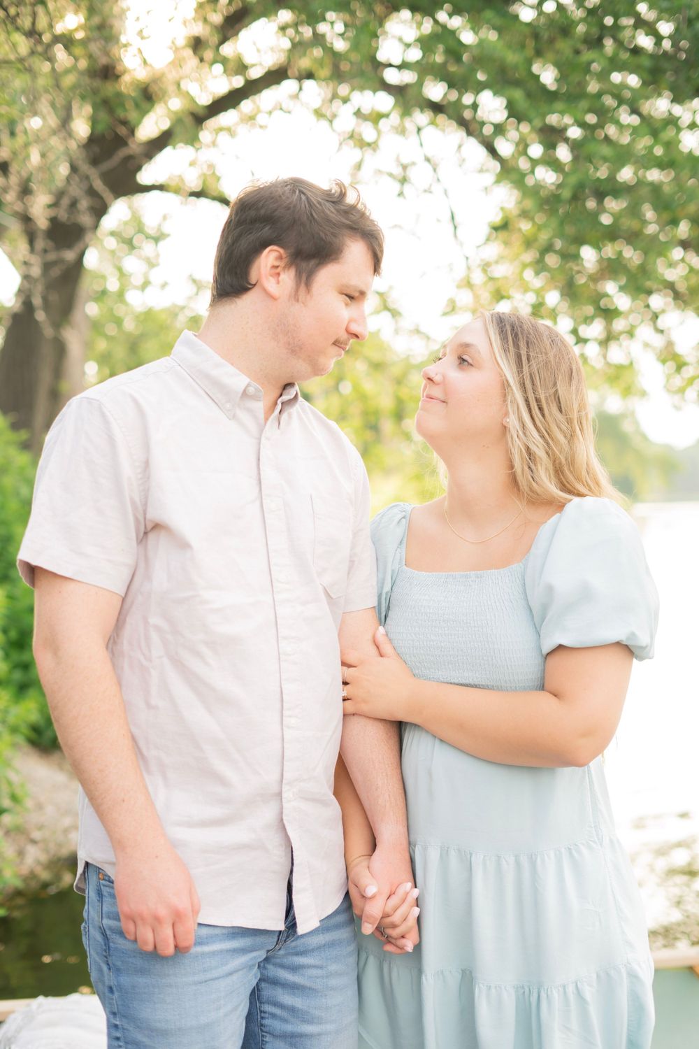 Couple gazing into each other's eyes while holding hands outdoors, surrounded by soft golden sunlight and green trees.