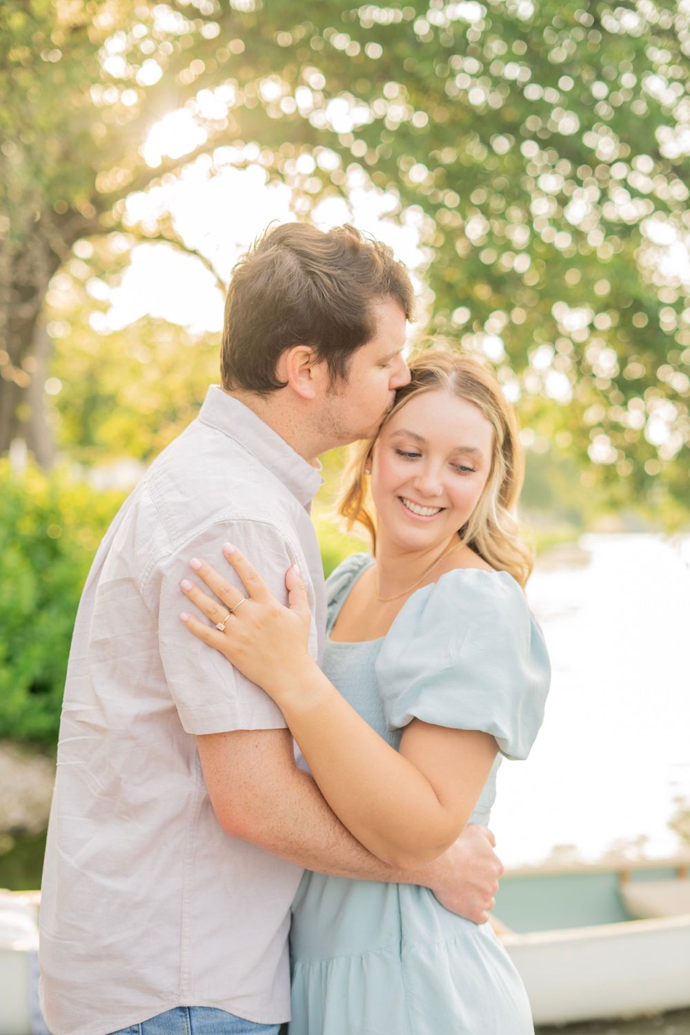 Couple embracing outdoors, man kissing woman's cheek as she smiles, golden sunlight filtering through trees near a lake.