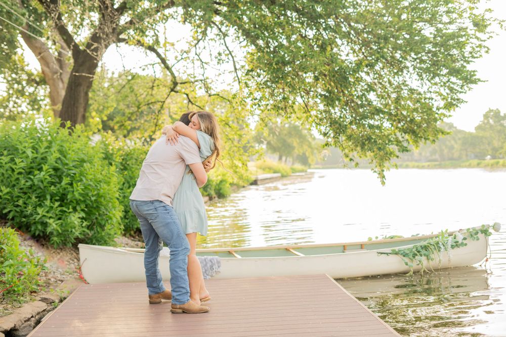 Couple embracing on a wooden dock beside a serene lake with a white canoe and green trees nearby.