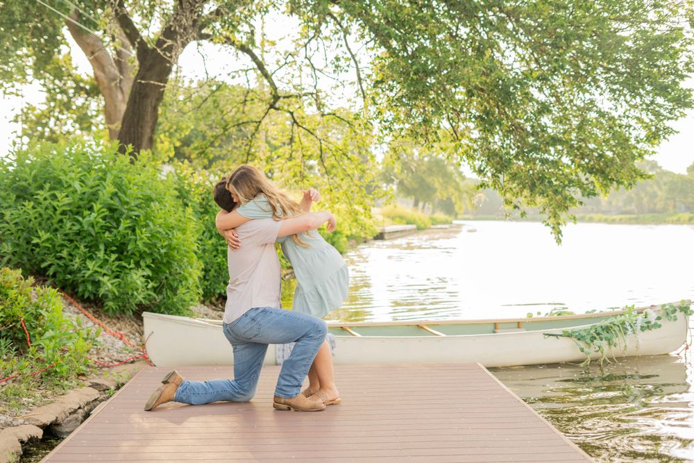 Man on one knee embracing his partner on a lakeside dock after a surprise marriage proposal.