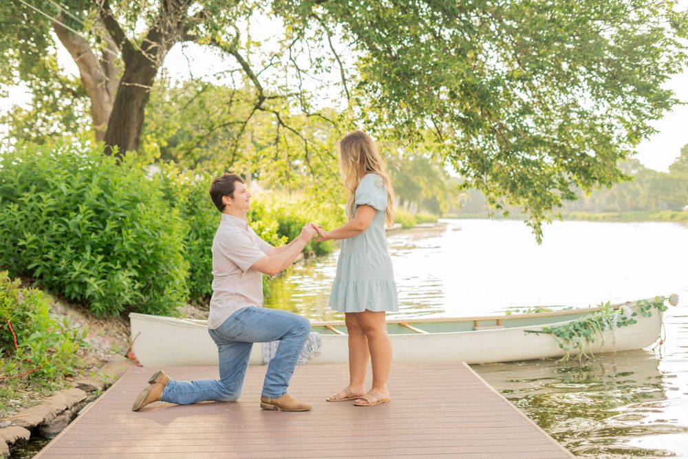 Man proposing marriage on one knee to his girlfriend on a scenic lakeside dock with a white rowboat.