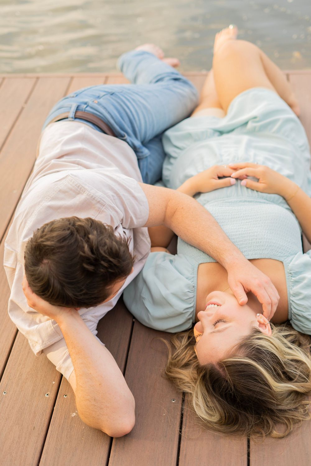 Couple lying on a wooden dock by the water, laughing and holding hands in a romantic lakeside moment.