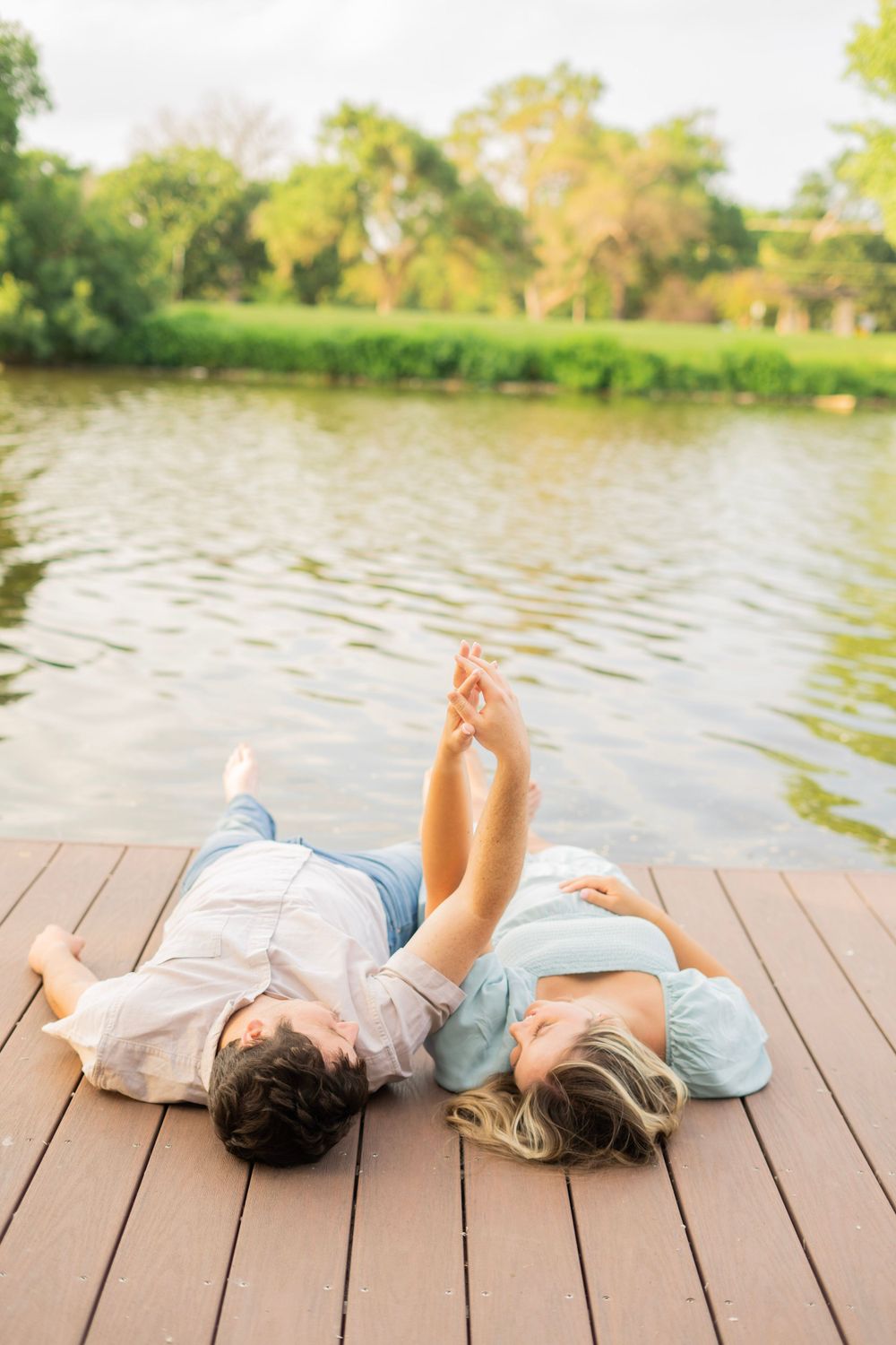 Couple lying on a dock with feet raised, holding hands playfully with a serene green lake backdrop.