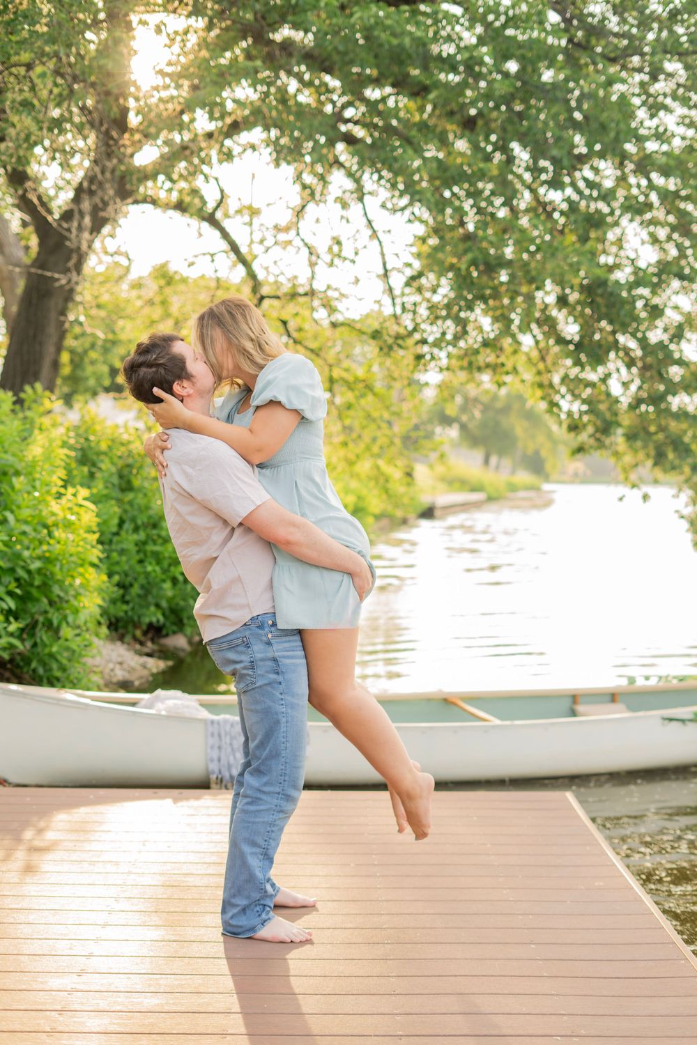 Man lifting and embracing woman on a sunny dock beside a white canoe by the river.