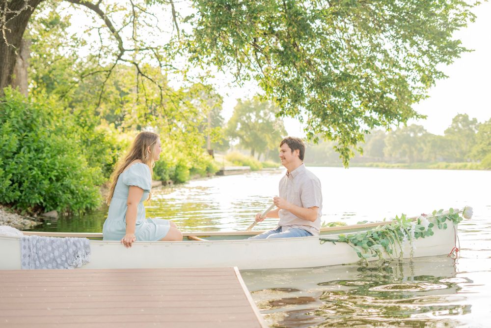 Happy couple sitting in a decorated white canoe on a calm lake surrounded by lush green trees.