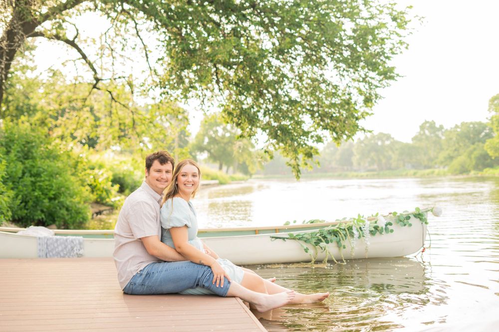 Smiling couple sitting together on a dock beside a flower-adorned white canoe with a peaceful lake backdrop.