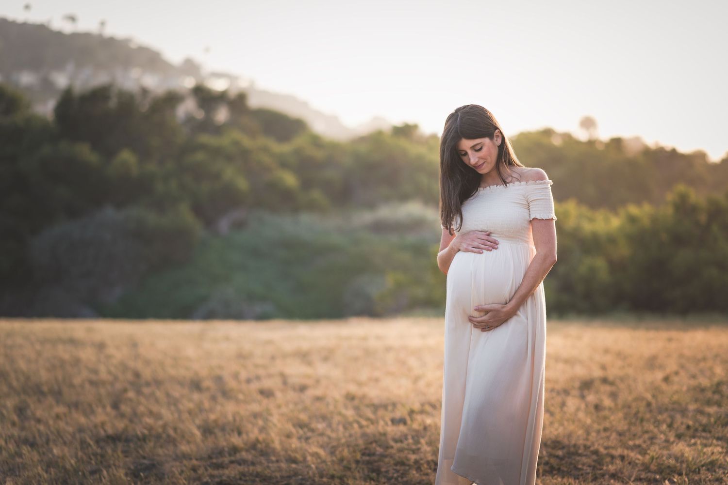 A pregnant woman in a white dress stands in a golden sunlit field at sunset.