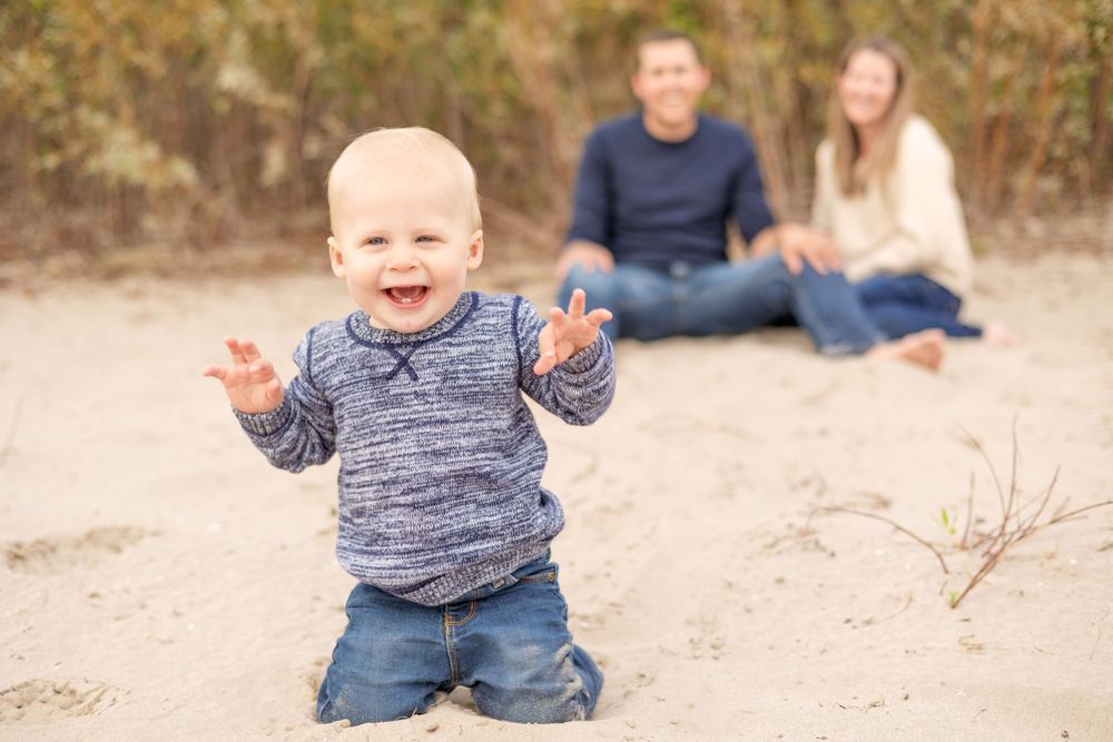 A happy toddler in gray sweater poses on sand with adults sitting in background.