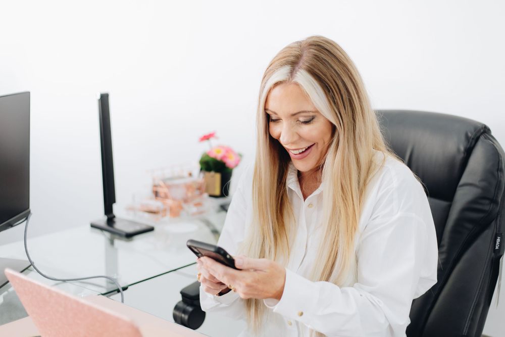 Person checking phone while sitting at office desk with pink laptop and flowers.