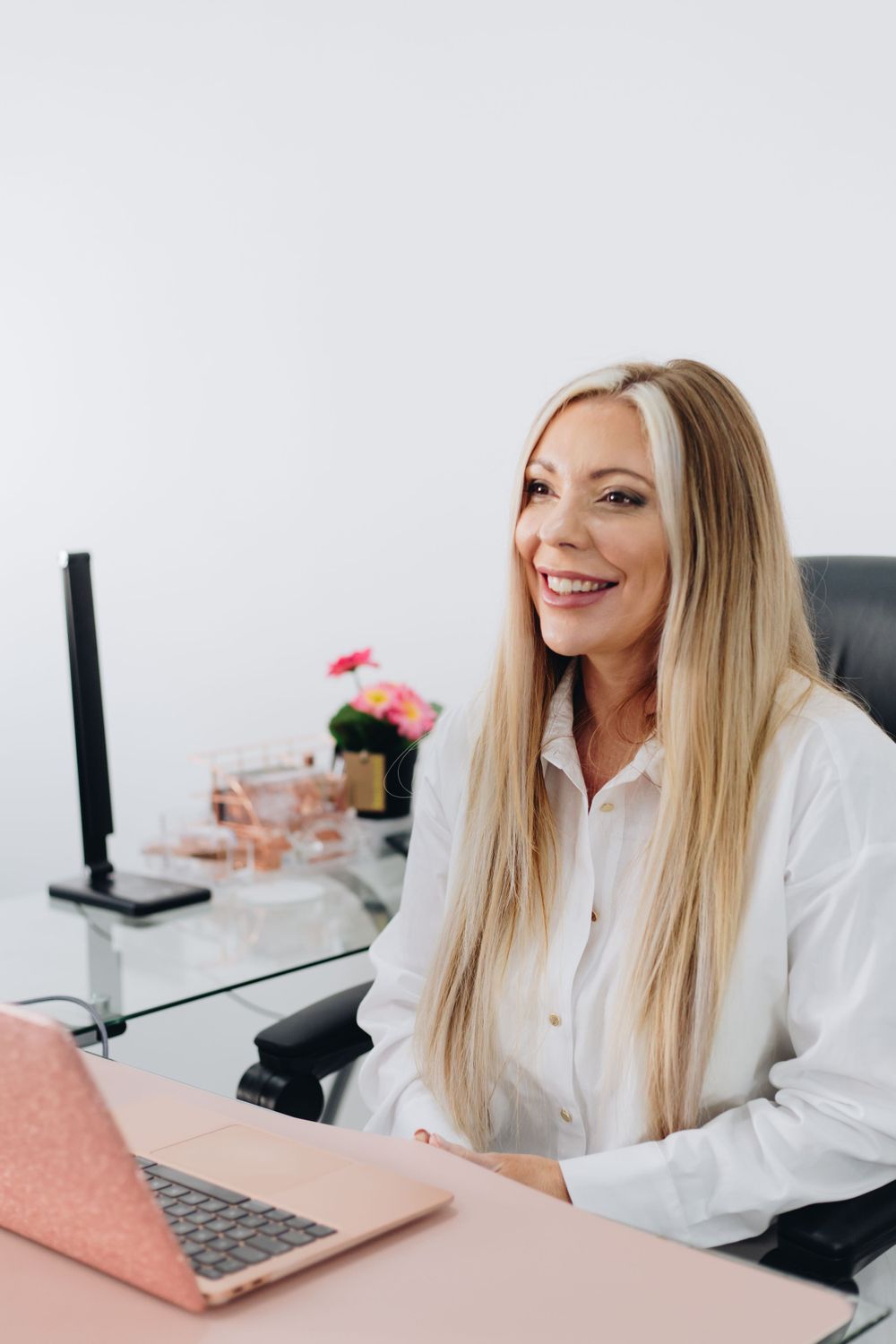 Business professional smiling at camera while working at pink desk with laptop and flowers.