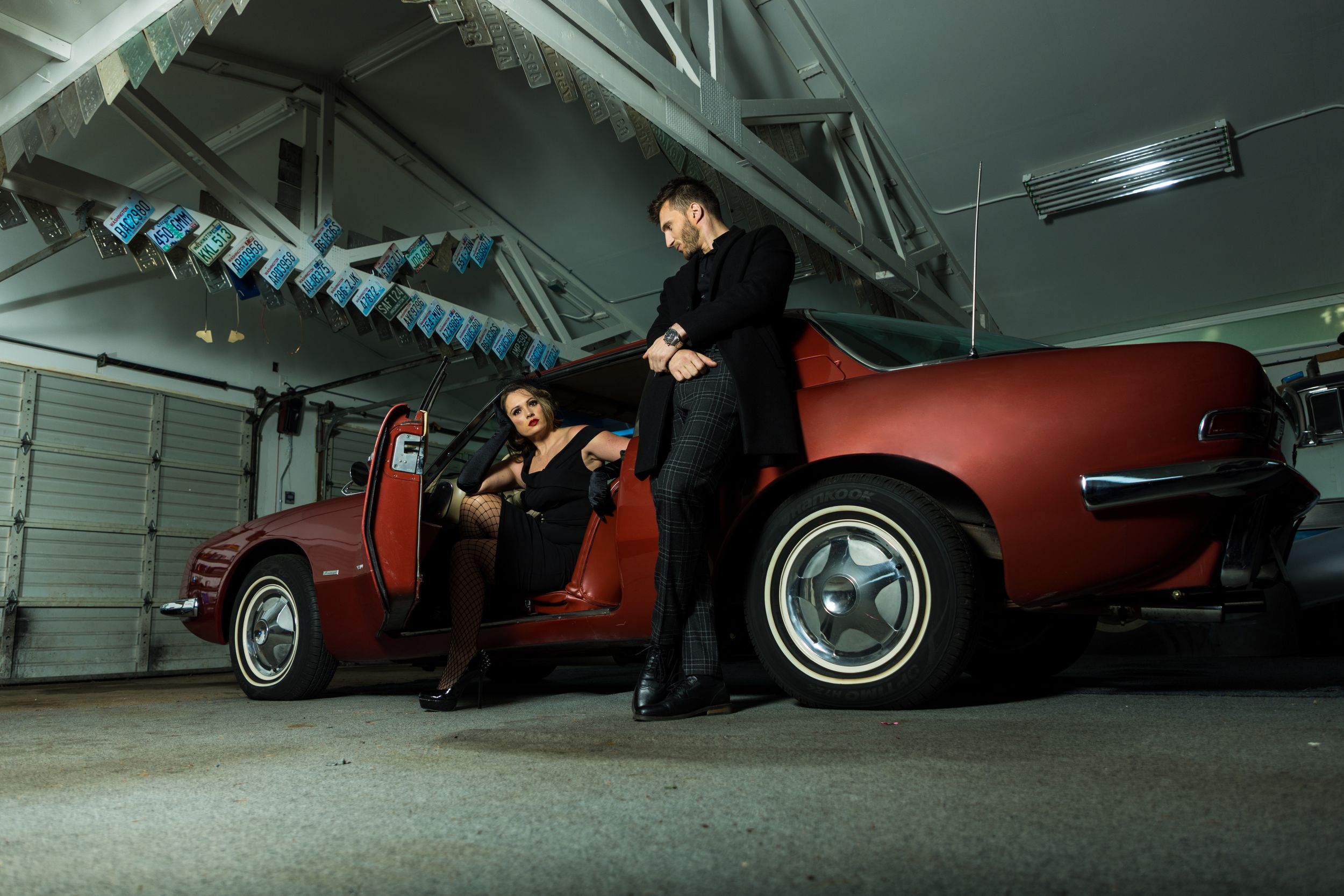 Group posing with classic red car in industrial garage with moody lighting.