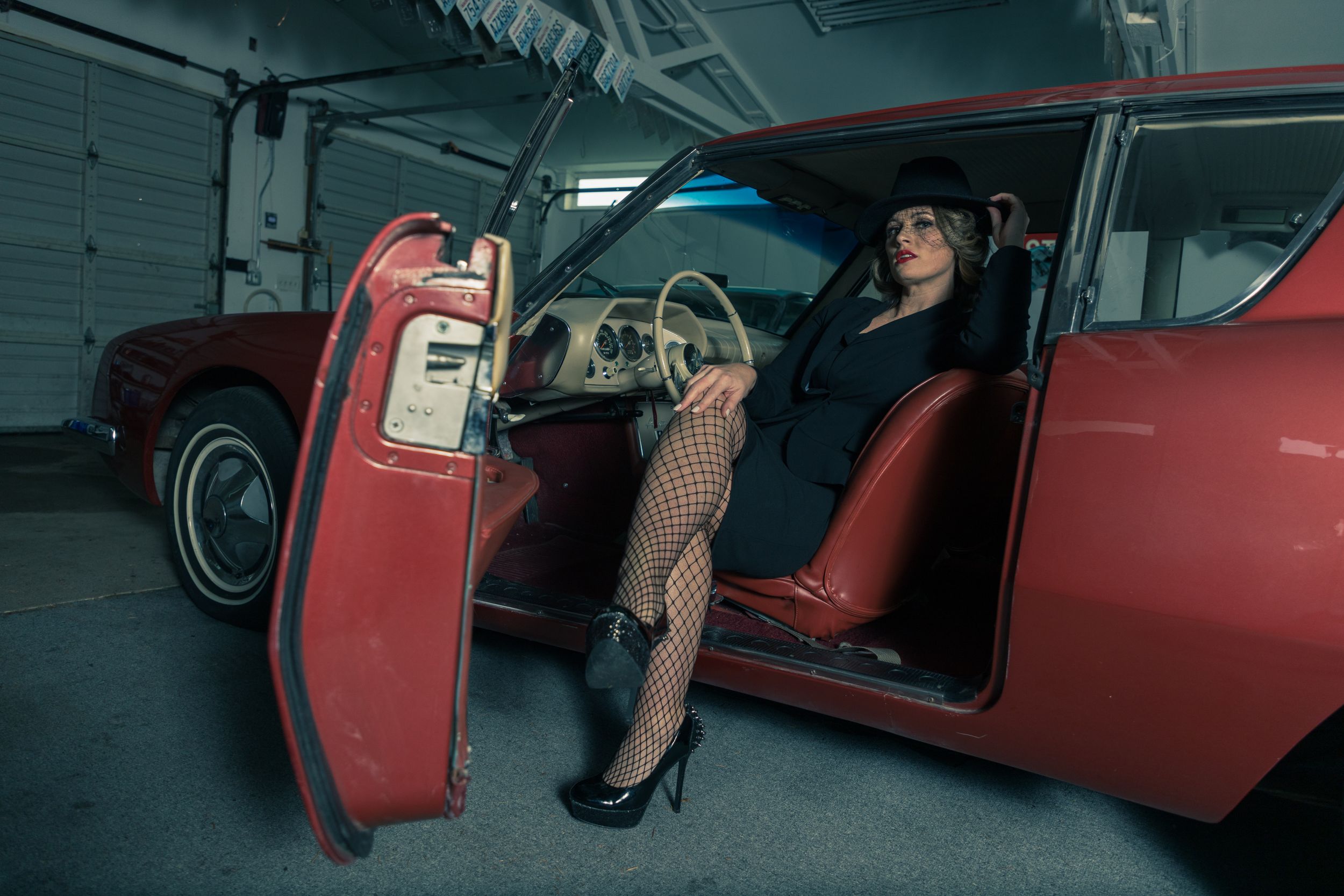 Woman in elegant attire sitting in red vintage car with door open in dimly lit garage.