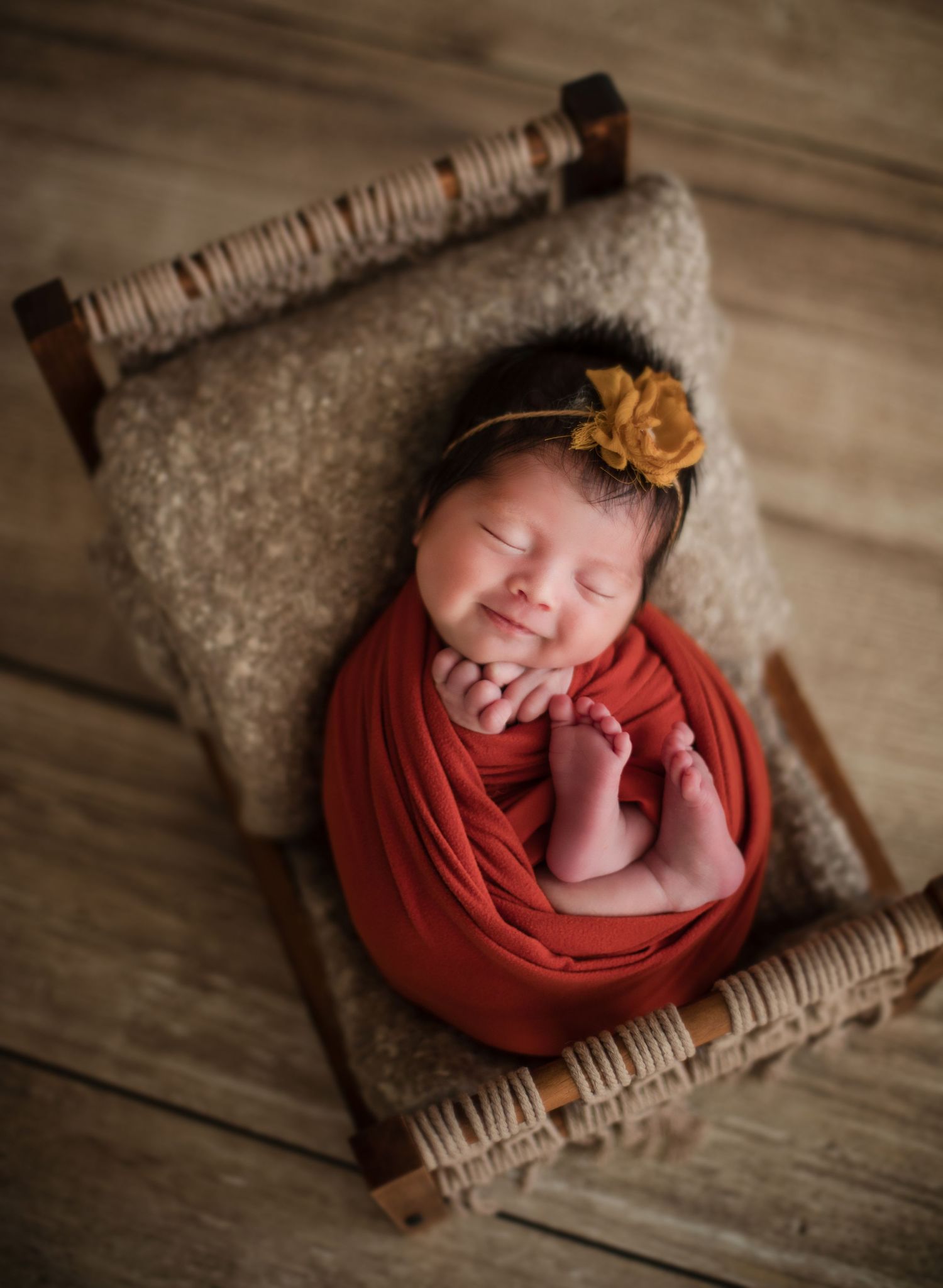 Smiling newborn baby swaddled in red wrap with yellow flower headband resting in rustic wooden bed.