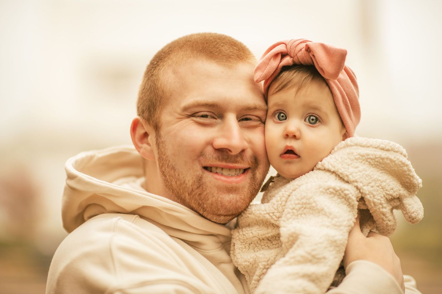 dad holding one year holding daughter touching cheeks. both looking and smiling at camera.
