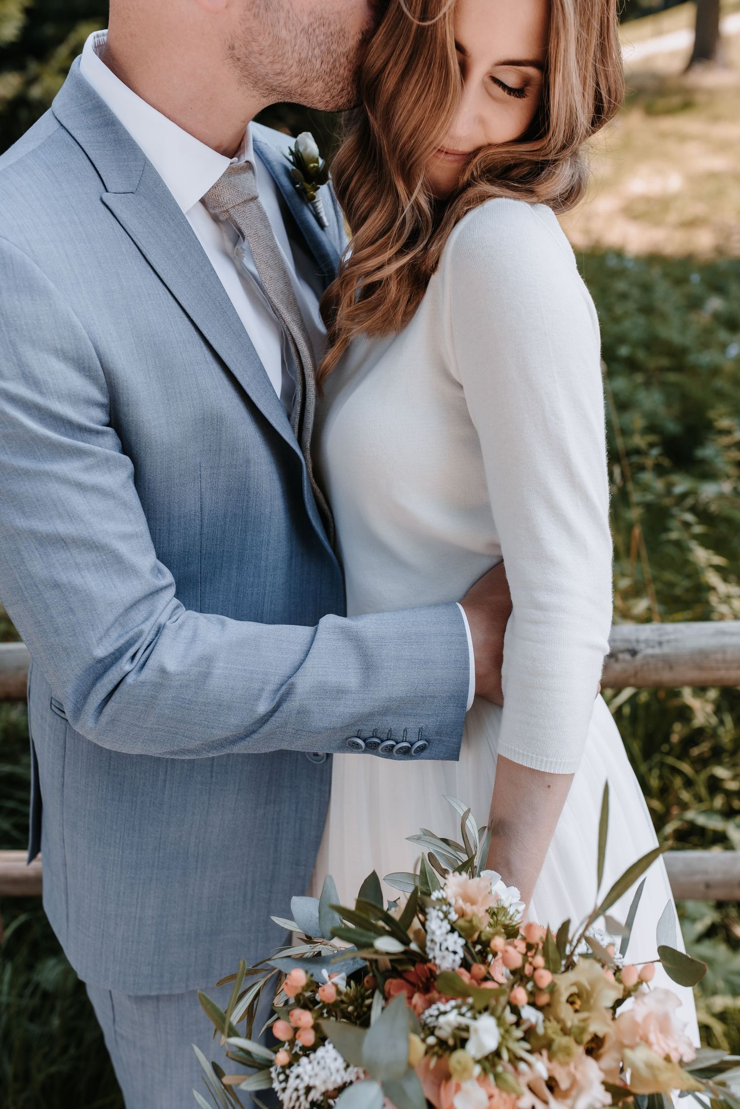 A romantic wedding portrait showing a couple embracing with a soft-focus floral bouquet in pastel tones.