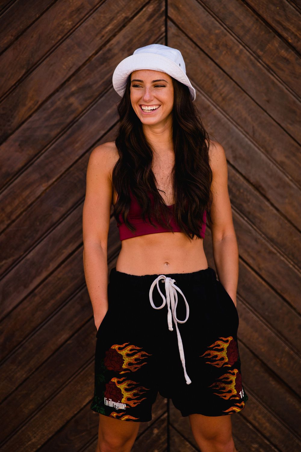 Casual streetwear pose featuring white bucket hat and flame-detailed shorts against wooden wall.