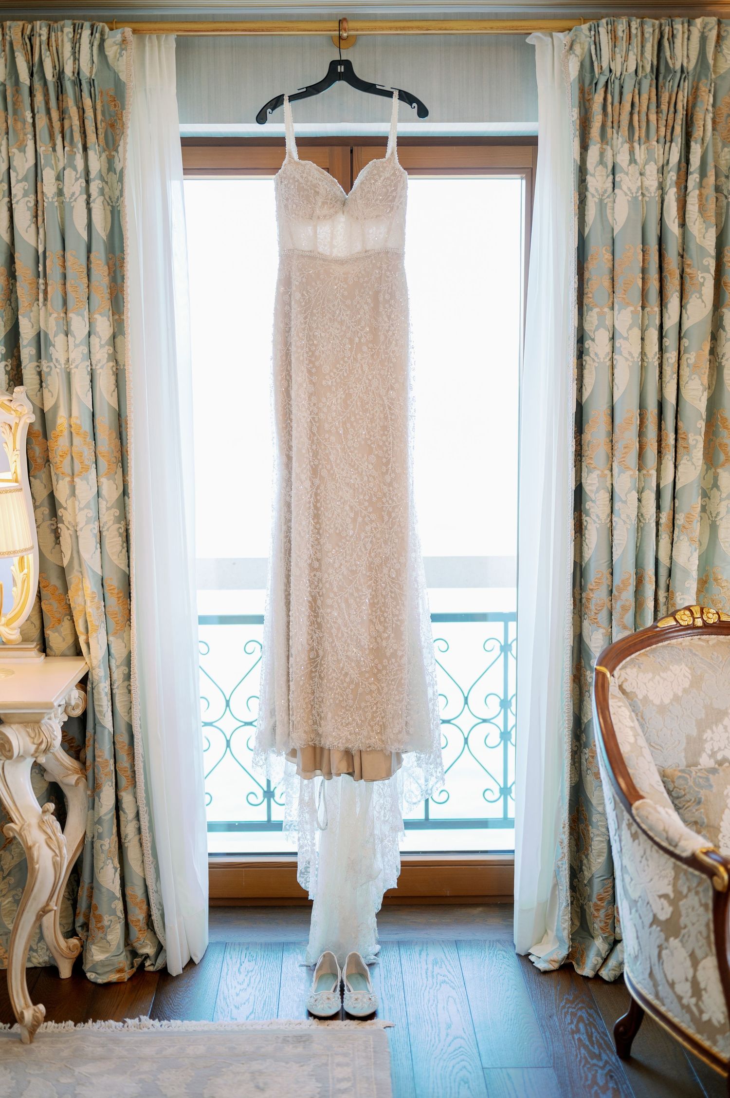 Blush wedding dress hanging in window with ornate curtains and French balcony.