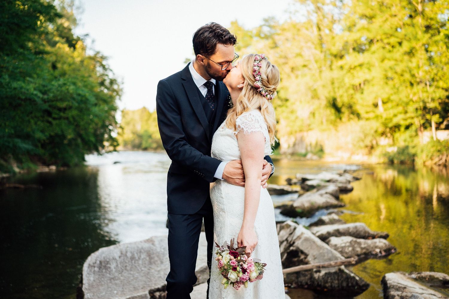 Couple in wedding attire embrace by lake with rocks and trees.