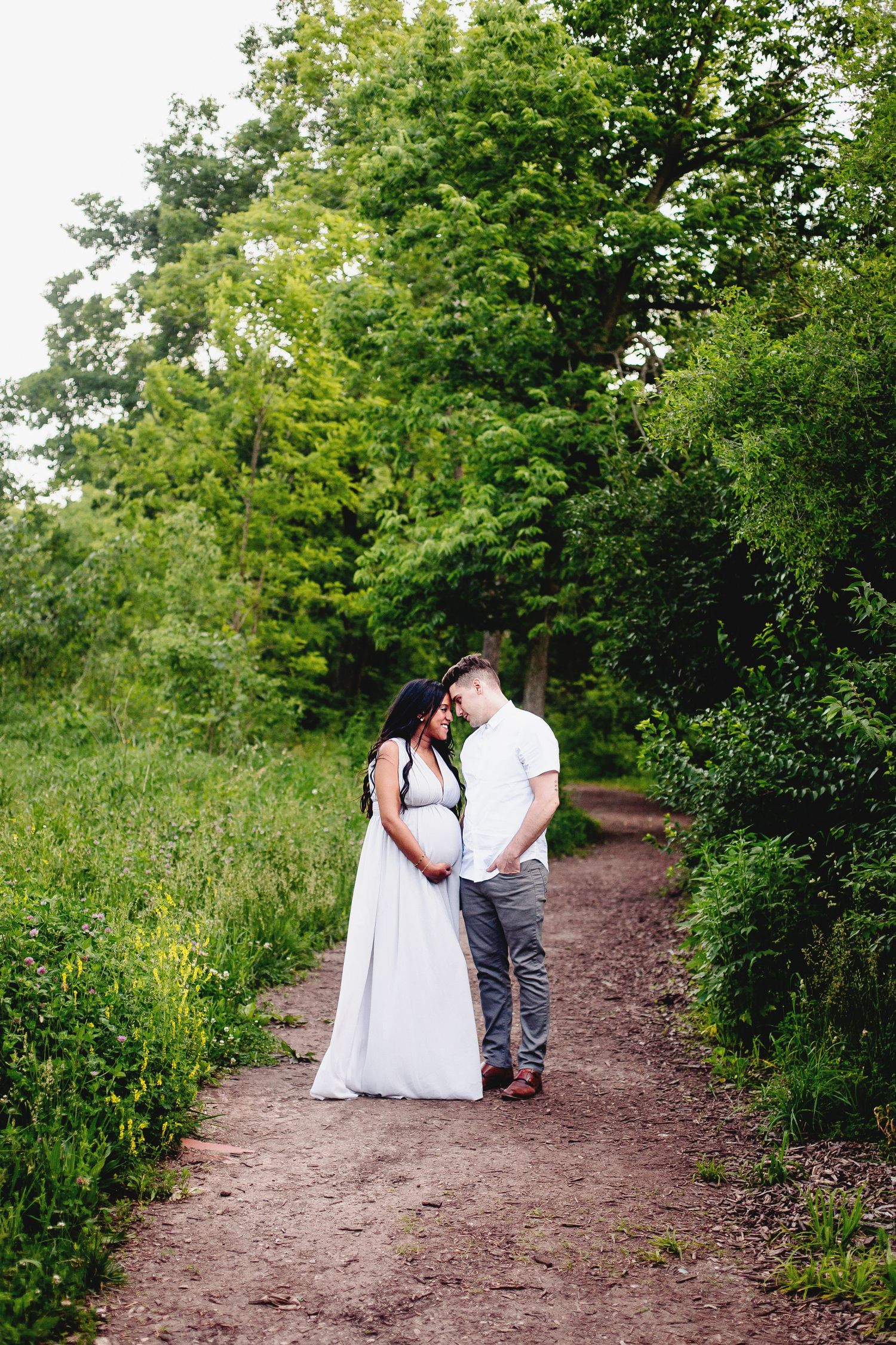 pregnant woman and man stand foreheads touching surround by green trees and bushes