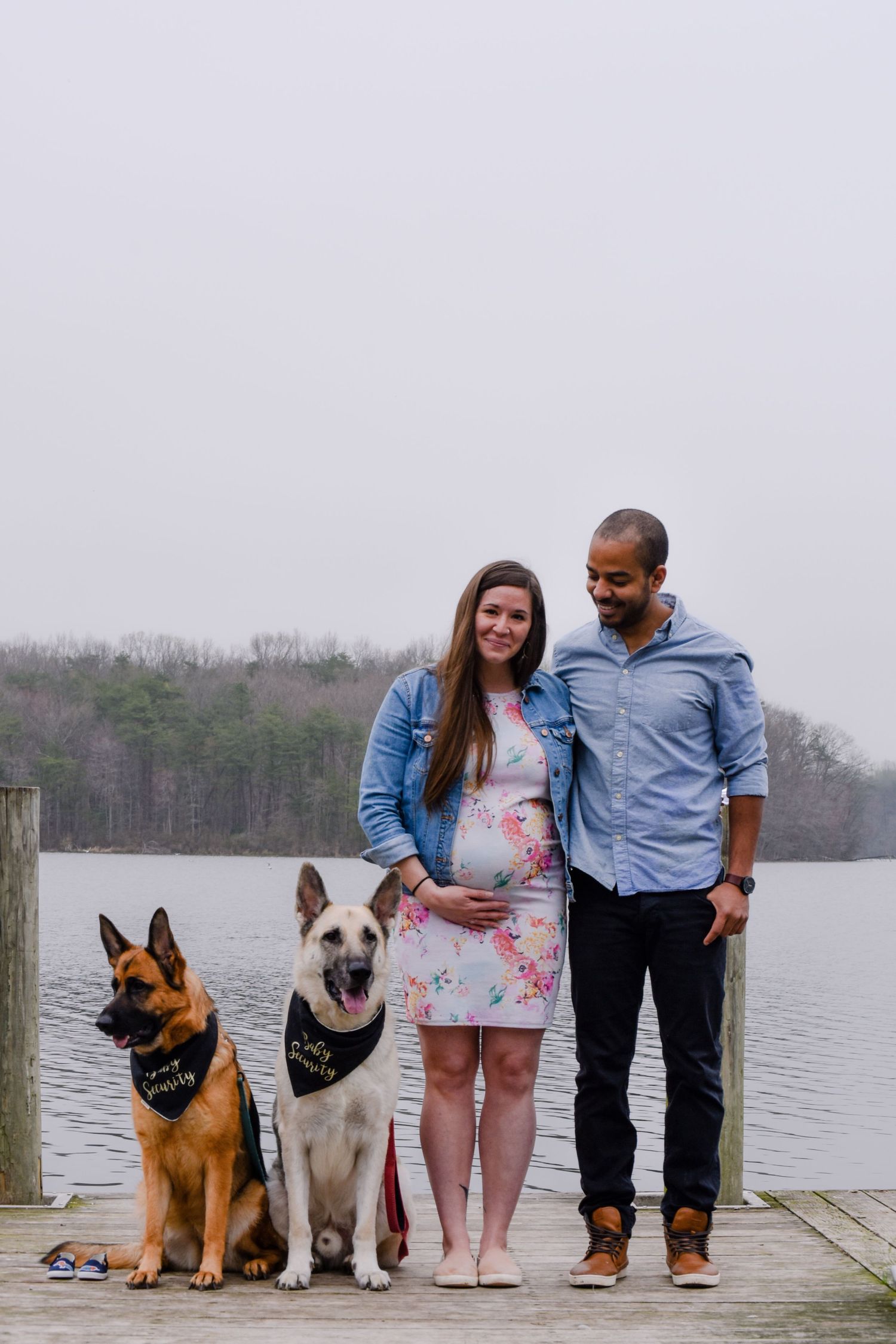 Couple with two dogs standing together on a dock by a lake with trees in the background.