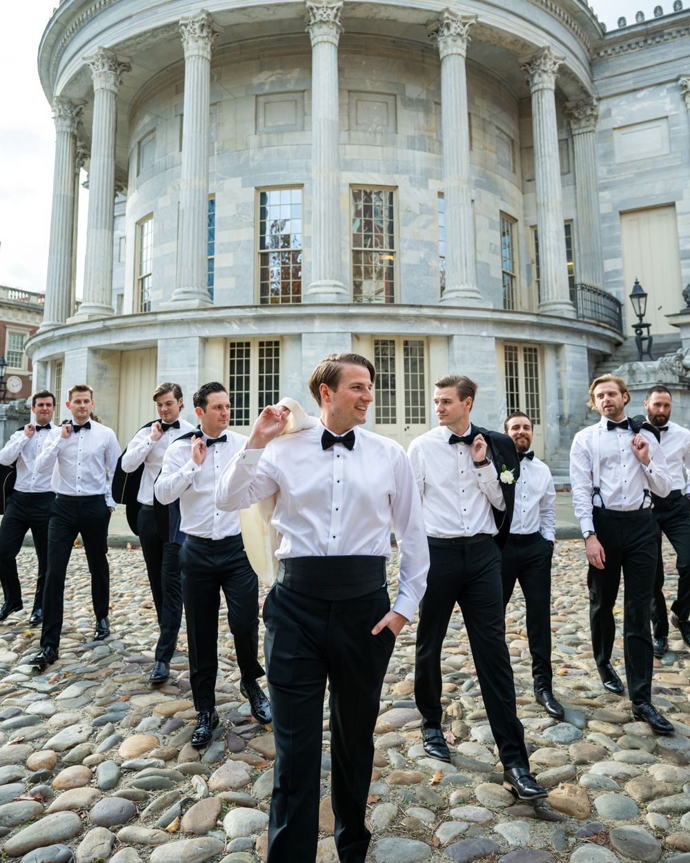 Groomsmen gather on cobblestones in front of a historic circular building with columns.