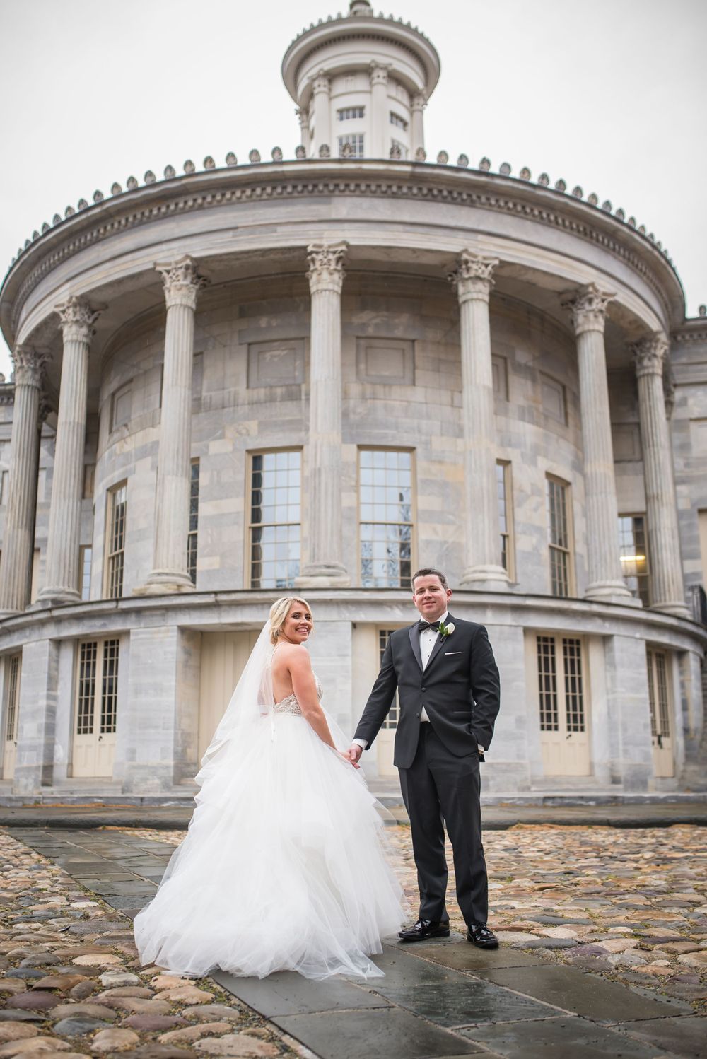 Wedding couple poses in front of a grand neoclassical building with white columns and circular architecture.