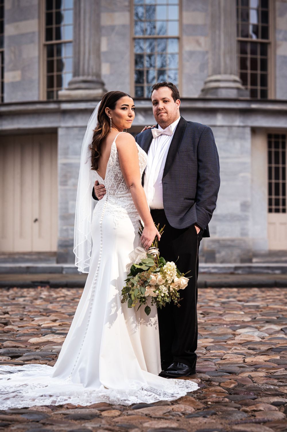 Wedding couple poses on cobblestone path with elegant stone building backdrop.