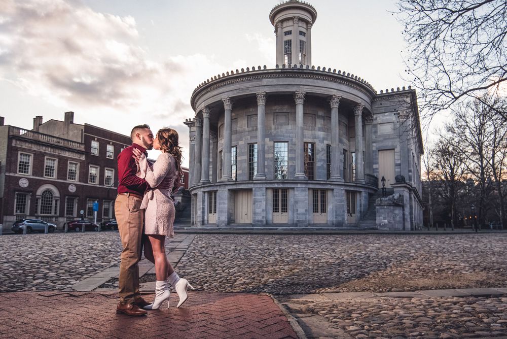 Casual engagement photo at sunset with historic circular building in background.