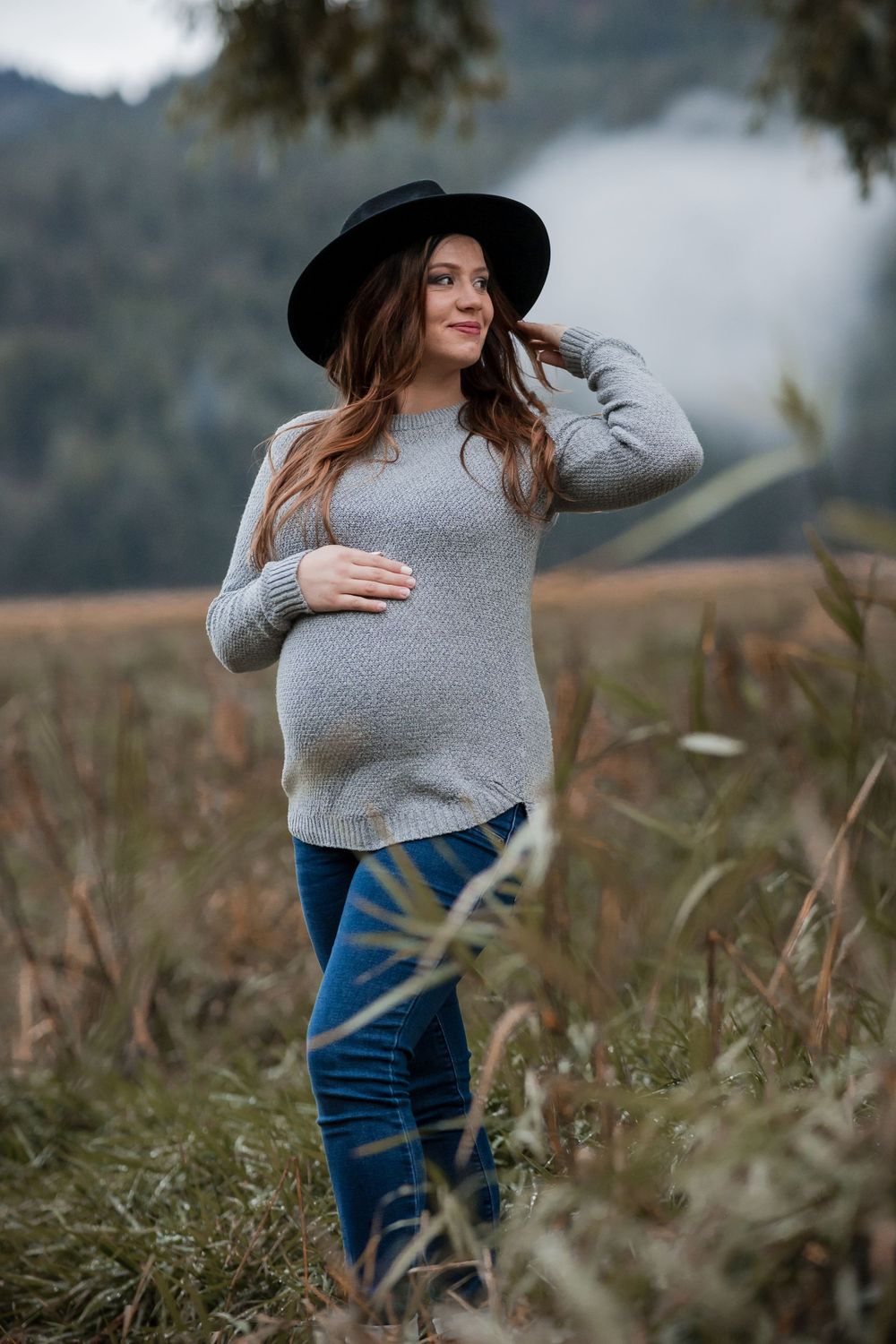 A pregnant woman in a grey shirt and blue jeans poses in long grass field during a maternity session in Liberty Lake.