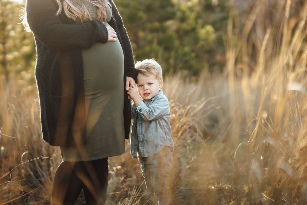 A pregnant woman and her young son stand together in tall grass during a maternity photo session.