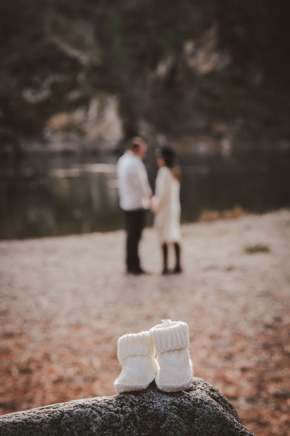 A pair of white baby shoes rests on a rock by the river, capturing a moment from a maternity session.