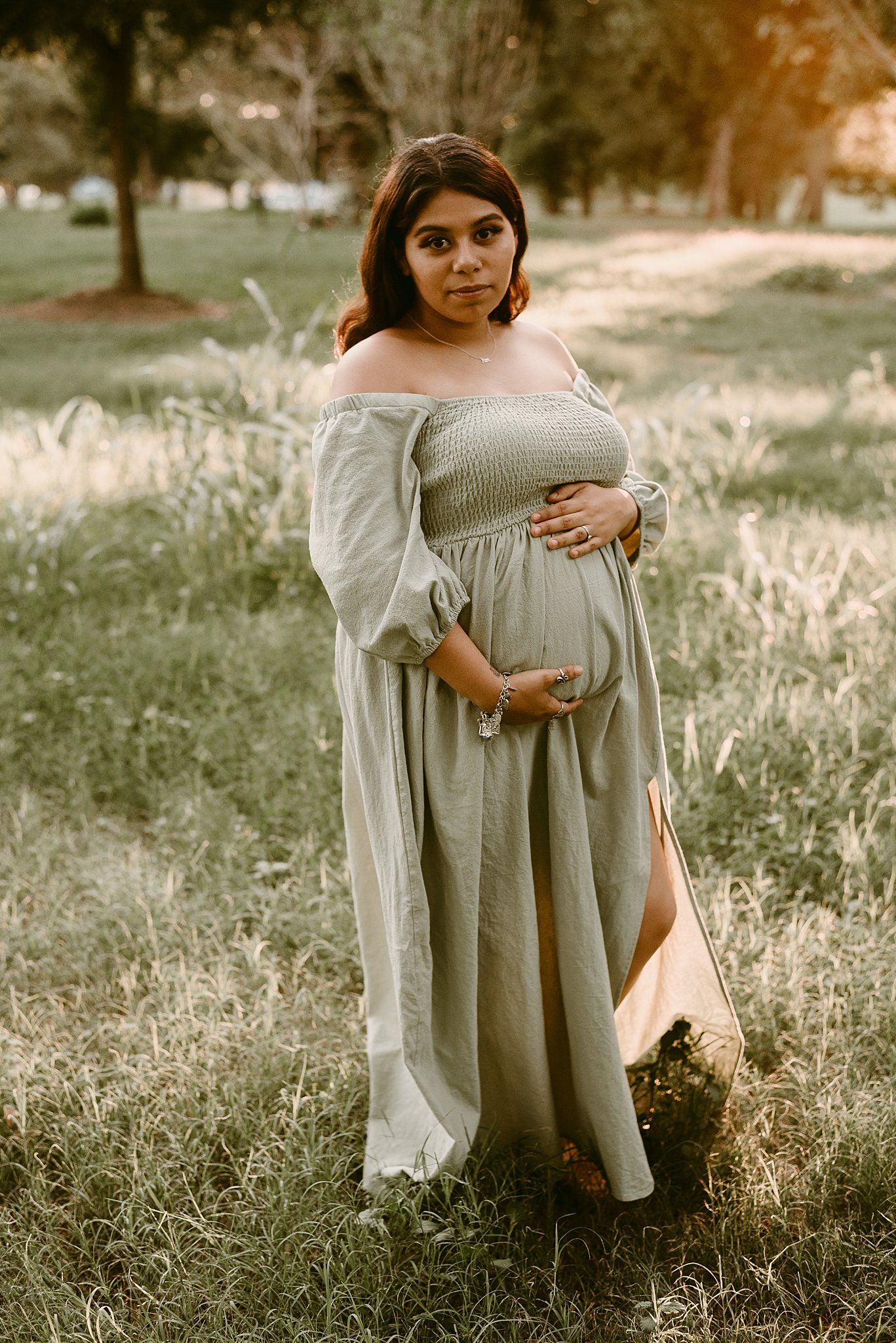 Maternity portrait of a person in an off-shoulder dress standing in sun-dappled grass.