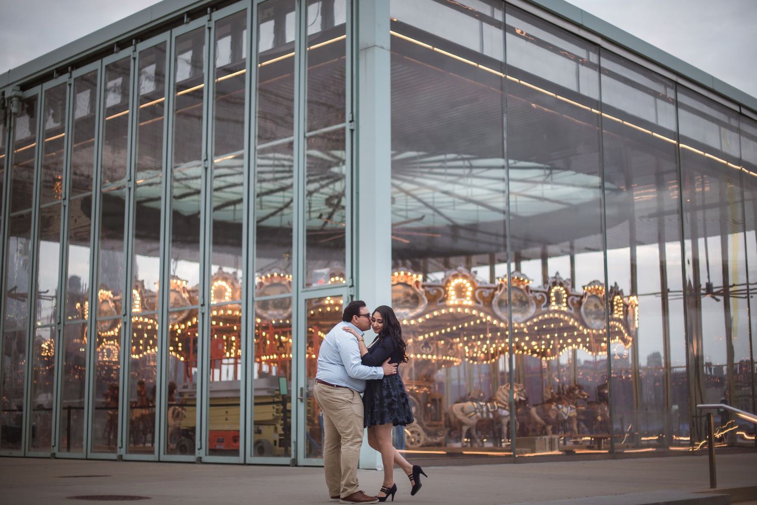 Couple sharing a romantic kiss in front of a glowing carousel at dusk during their engagement session.
