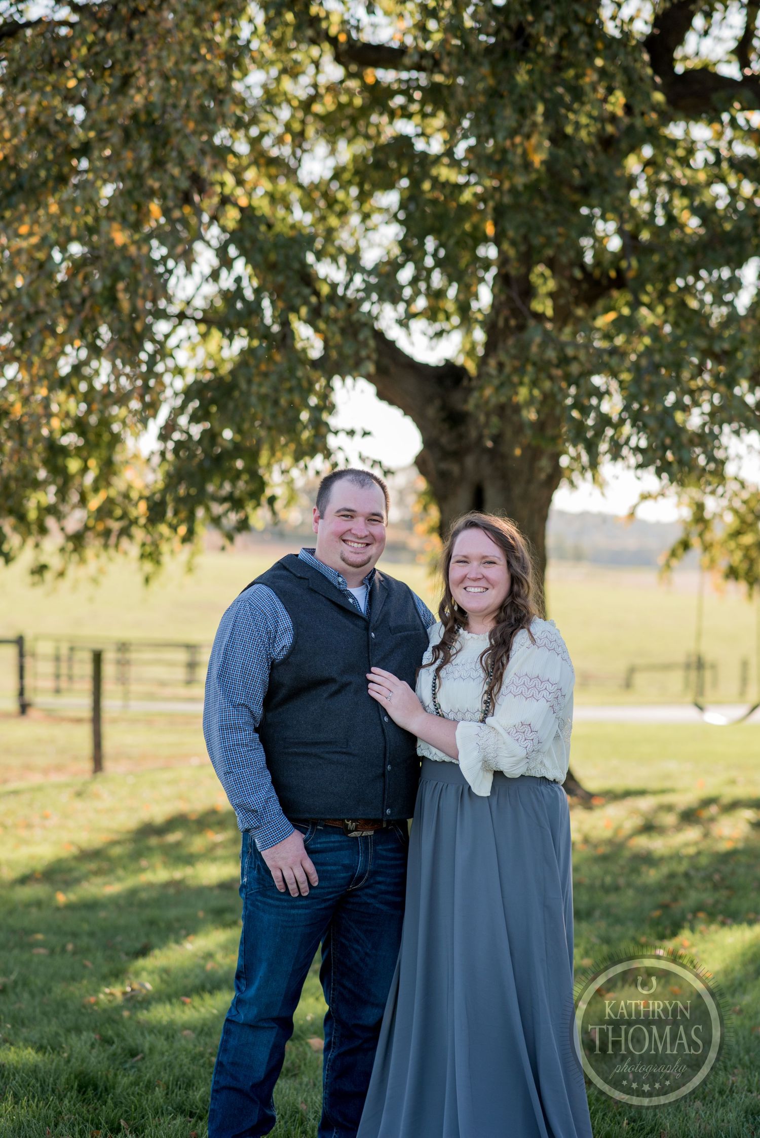 Couple poses together under large tree branch in sunny outdoor setting.