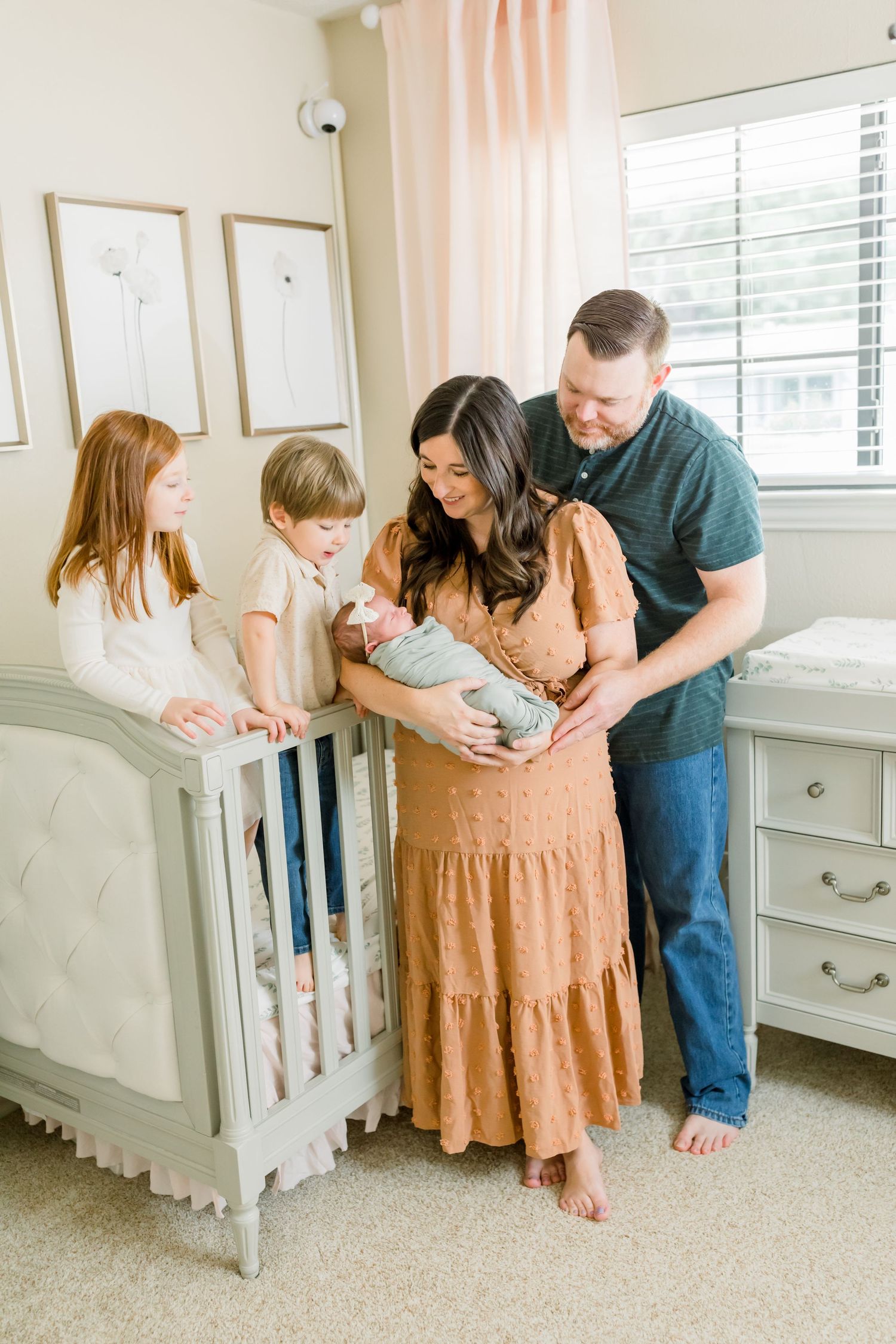A family gathers around a gray crib in a nursery with peach curtains and wall art.
