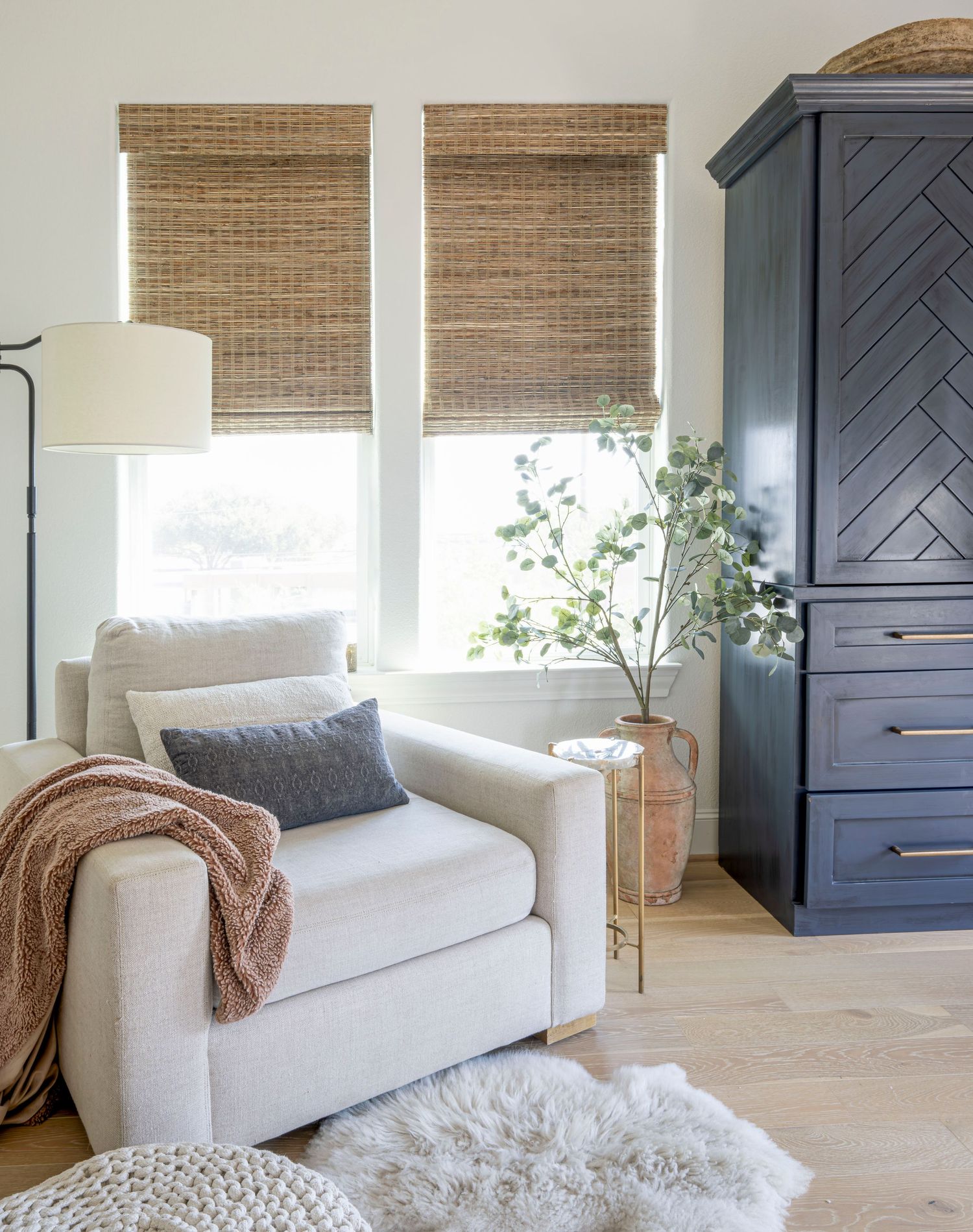 Modern living room corner with white armchair, bamboo shades and navy blue storage cabinet.