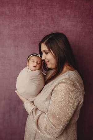 Mother in sparkly beige dress holding newborn against burgundy backdrop in studio setting.