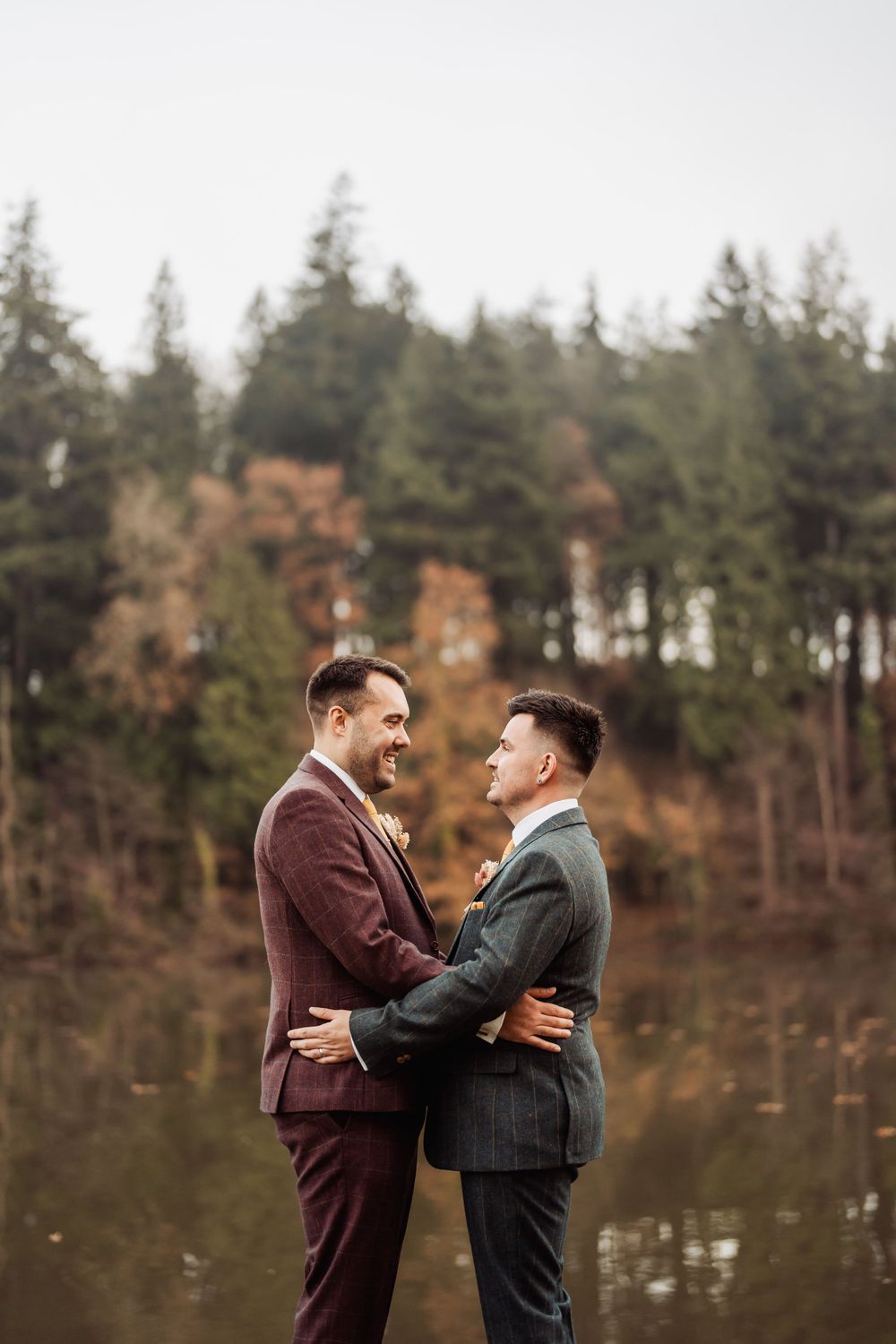 Two people in formal suits embrace tenderly by a serene lake surrounded by evergreen trees in an autumn setting.