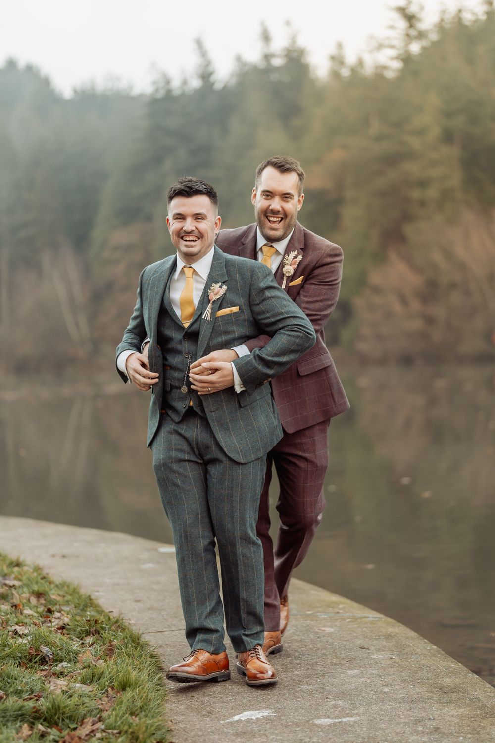 Two people in coordinated suits share a playful moment on a pathway by a misty lake.