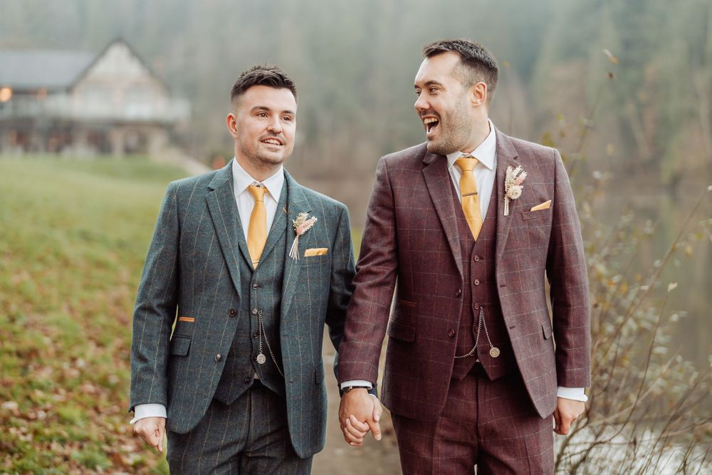 A couple in burgundy and gray suits walks hand-in-hand through a scenic countryside setting.
